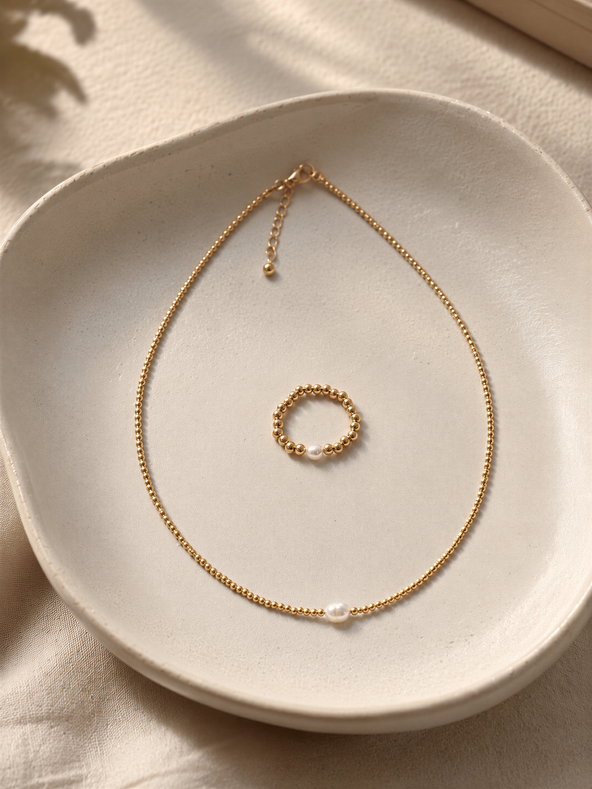 A delicate gold necklace with a round pendant and a small round ring are displayed on a white plate.