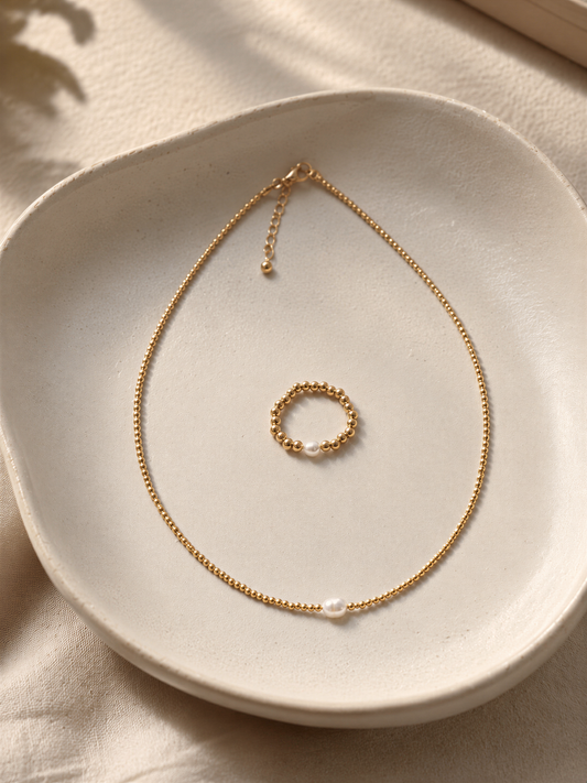 A delicate gold necklace with a round pendant and a small round ring are displayed on a white plate.