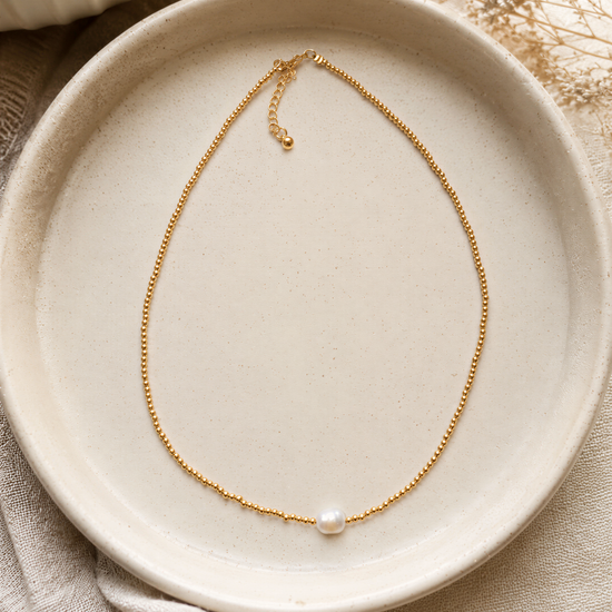 A delicate gold necklace with a single pearl pendant rests on a white plate, surrounded by a beige cloth and dried flowers.