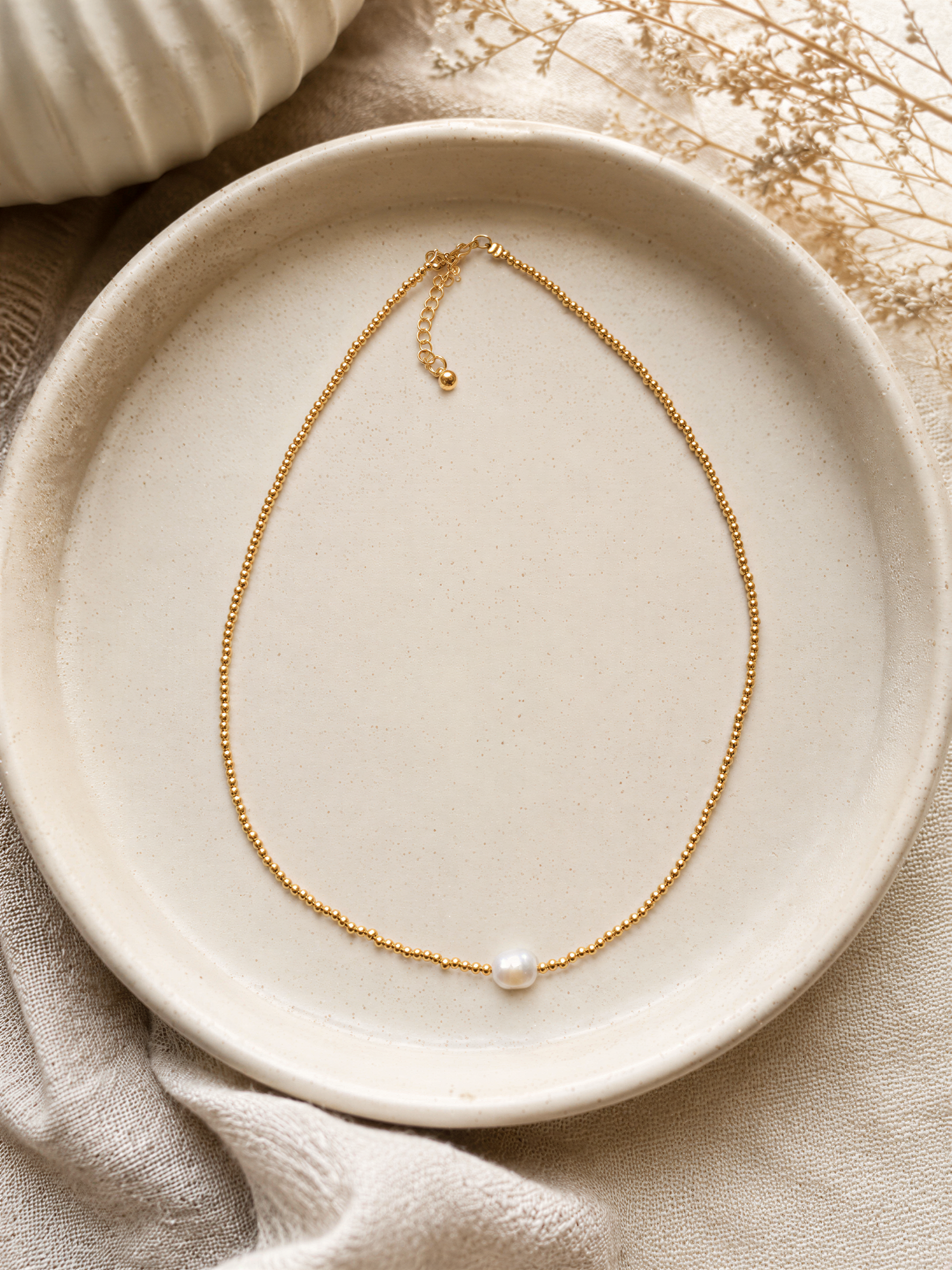 A delicate gold necklace with a single pearl pendant rests on a white plate, surrounded by a beige cloth and dried flowers.