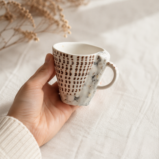 A hand holding a white mug with a patterned design, against a blurred background of dried flowers.