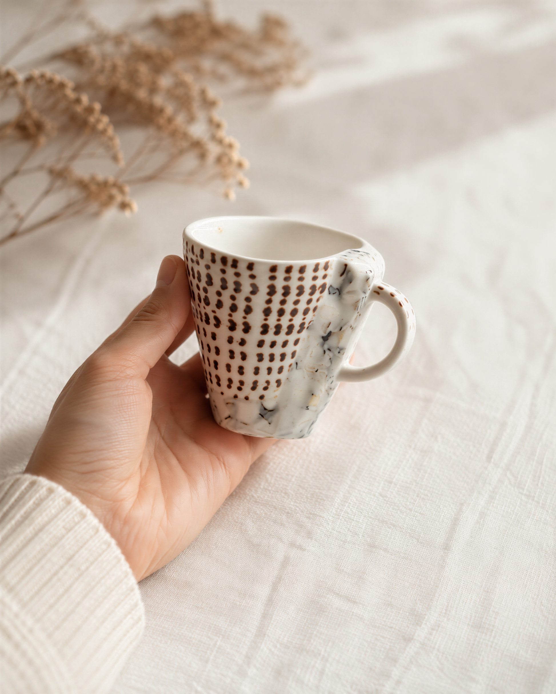 A hand holding a white mug with a patterned design, against a blurred background of dried flowers.