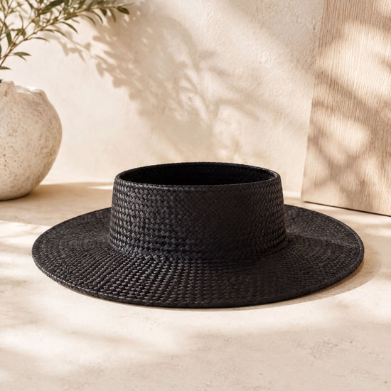 A black woven hat sits on a light-colored surface, with a white vase and a plant in the background.