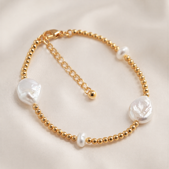 A gold-colored bracelet with three white pearls and a clasp, resting on a white fabric background.