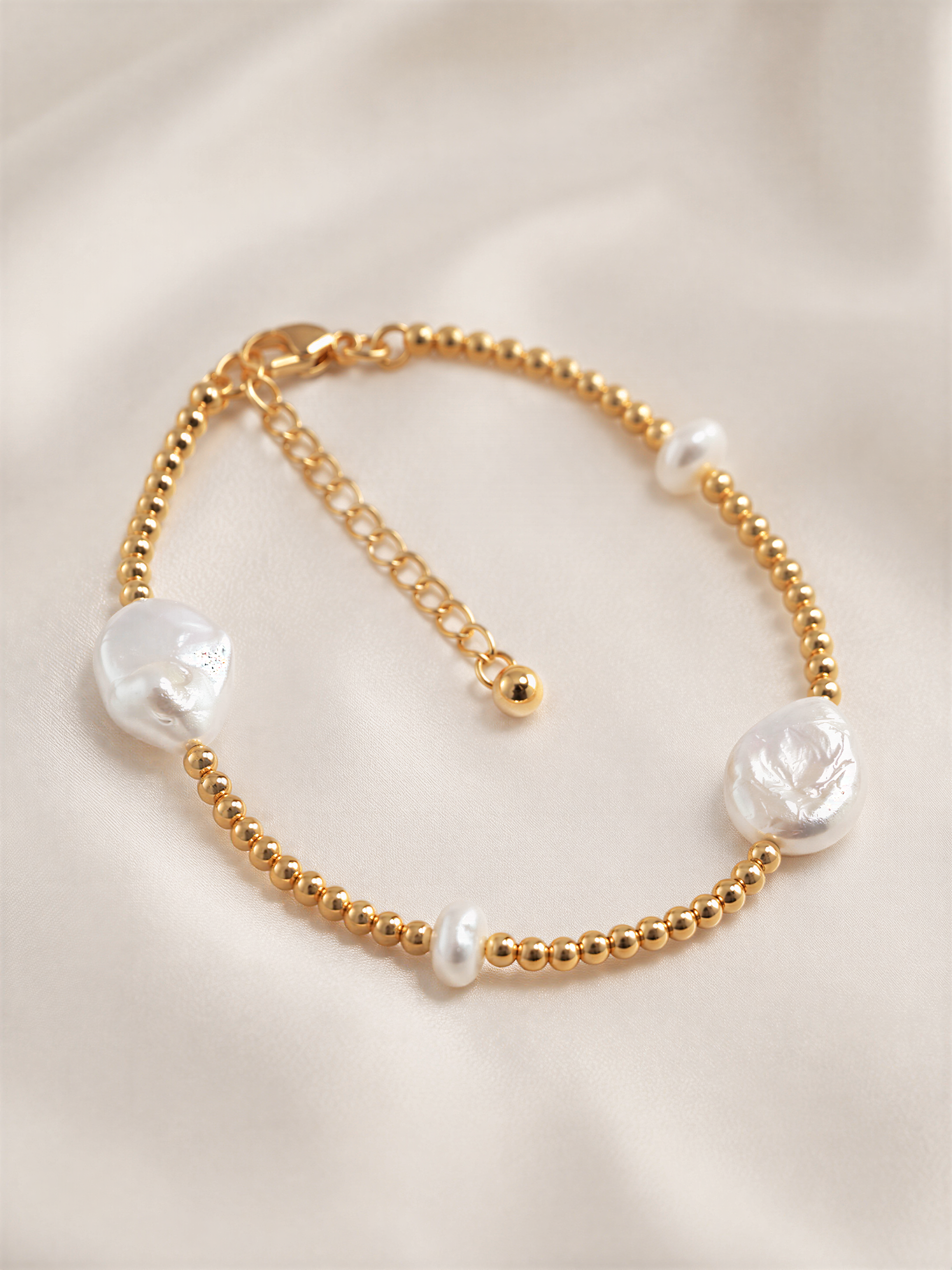 A gold-colored bracelet with three white pearls and a clasp, resting on a white fabric background.