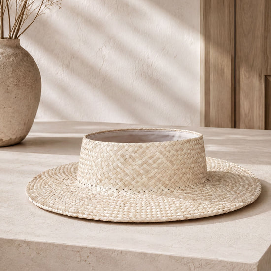 a straw hat placed on a white surface, with a vase of dried flowers to the left.
