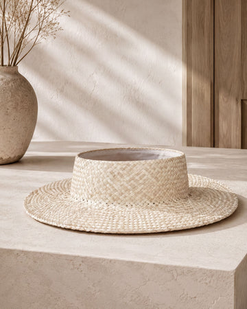 a straw hat placed on a white surface, with a vase of dried flowers to the left.