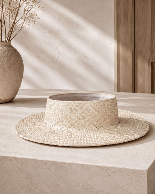 a straw hat placed on a white surface, with a vase of dried flowers to the left.