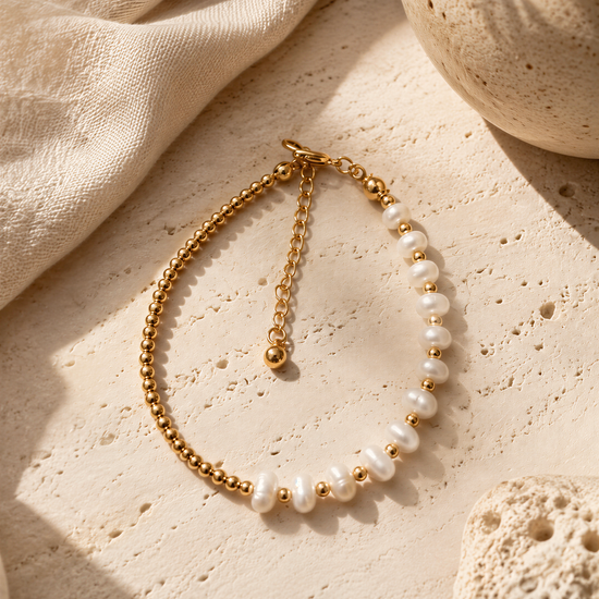 A gold-toned necklace with white pearls is displayed on a beige surface, accompanied by a small white vase and a piece of coral.