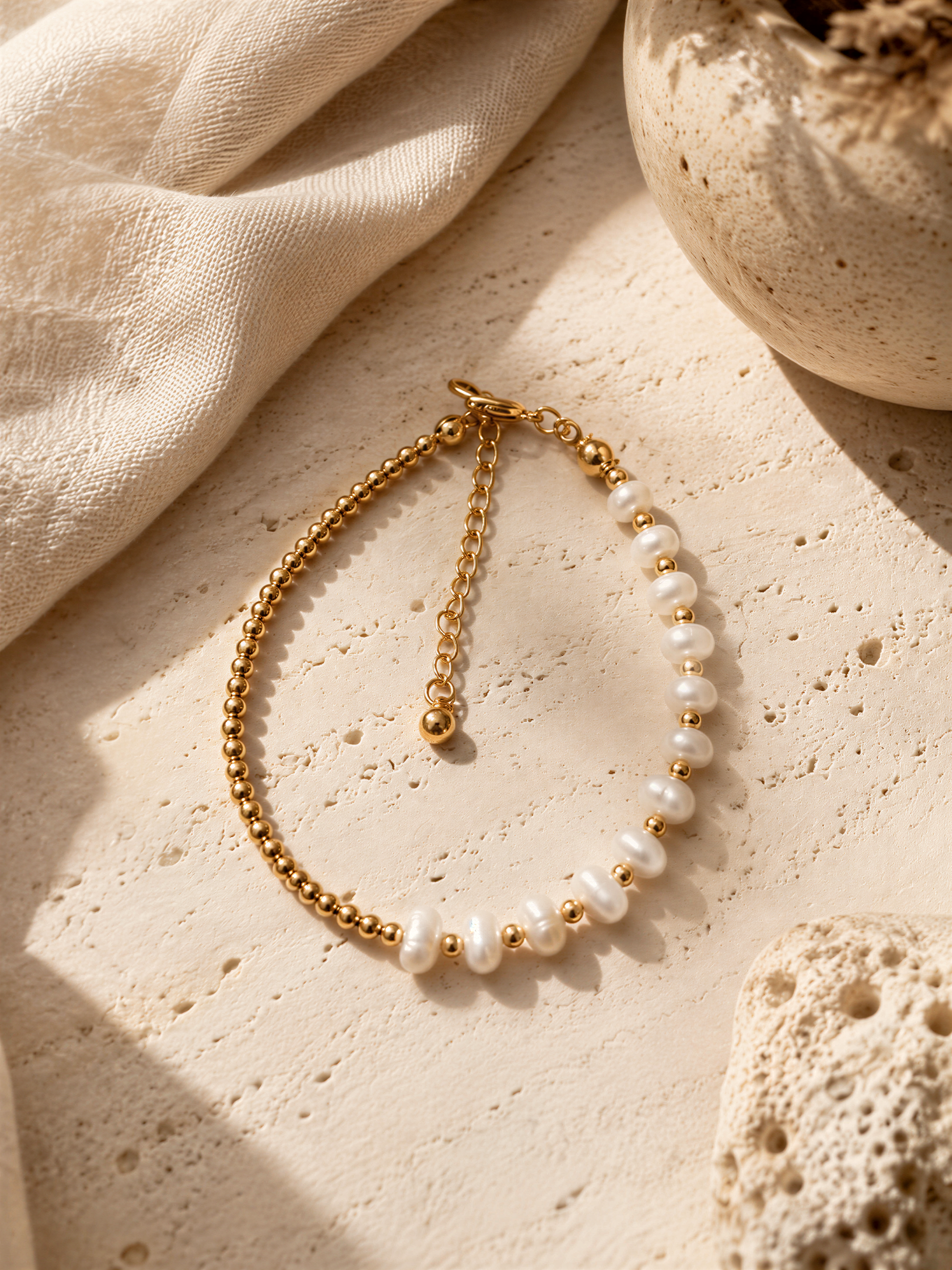 A gold-toned necklace with white pearls is displayed on a beige surface, accompanied by a small white vase and a piece of coral.