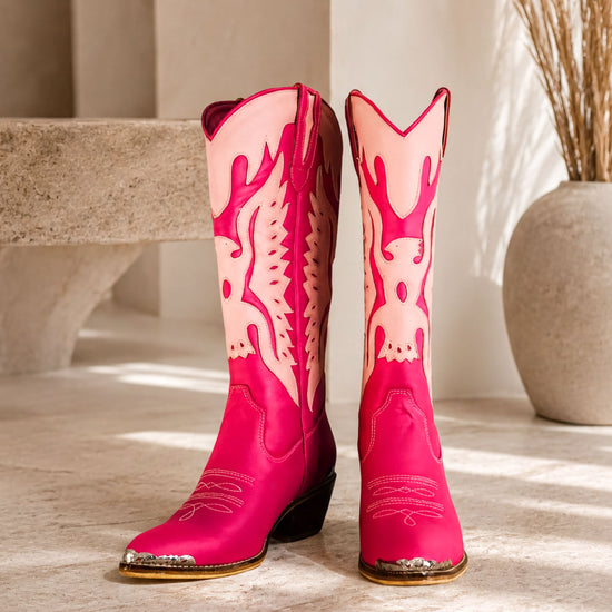 A pair of bright pink cowboy boots with white flames on the sides, placed on a white surface against a beige wall.