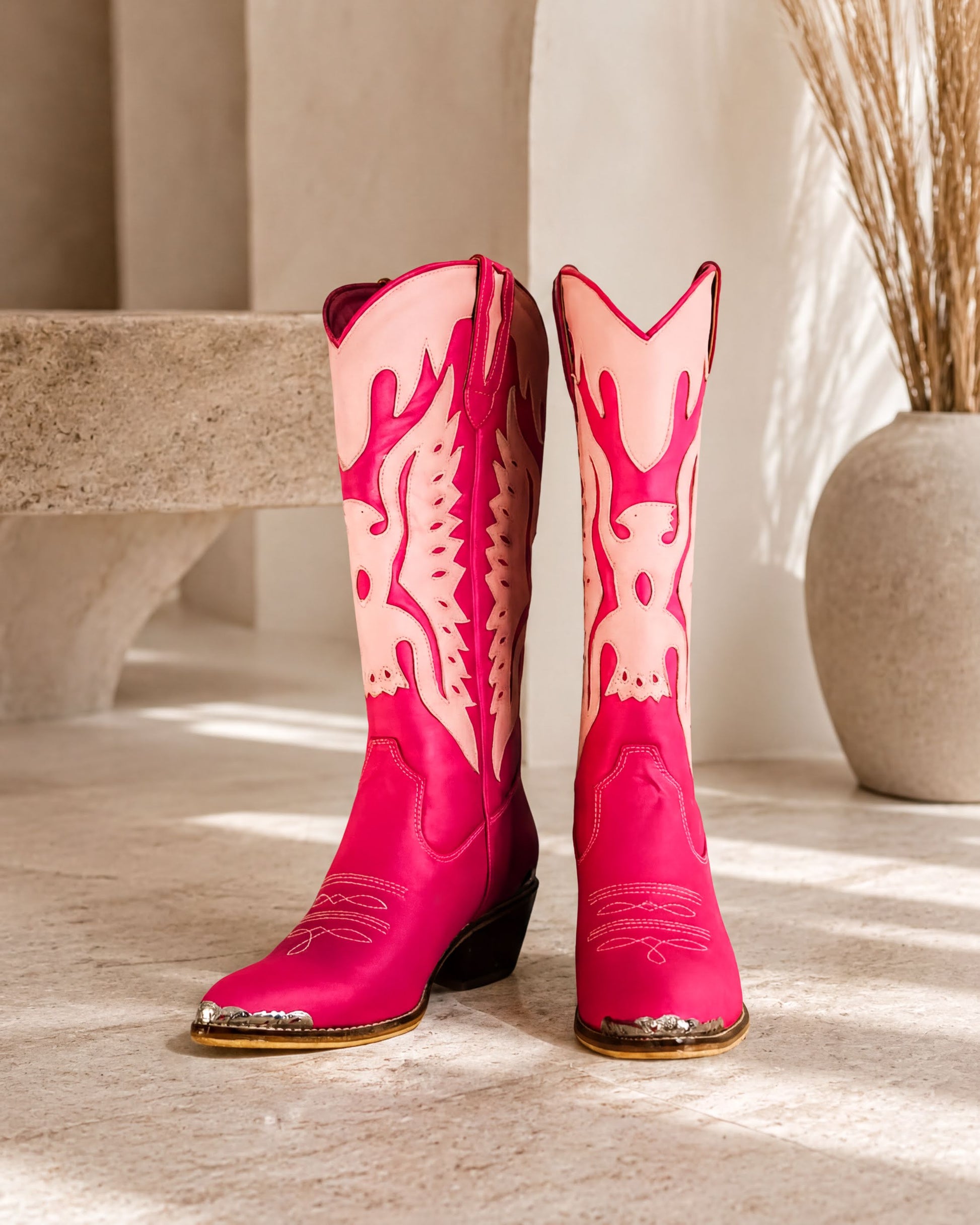 A pair of bright pink cowboy boots with white flames on the sides, placed on a white surface against a beige wall.