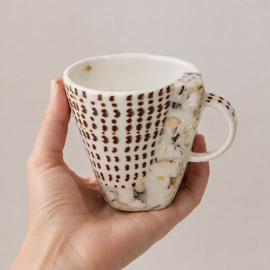 A hand holding a white mug with a brown patterned design.