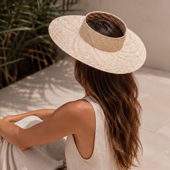 A woman wearing a wide-brimmed hat sits on a tiled floor, her back to the camera, with a lush green plant in the background.
