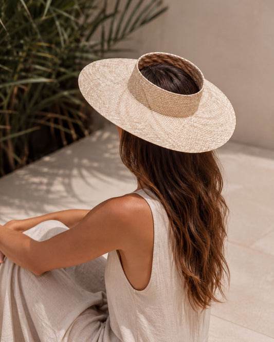 A woman wearing a wide-brimmed hat sits on a tiled floor, her back to the camera, with a lush green plant in the background.