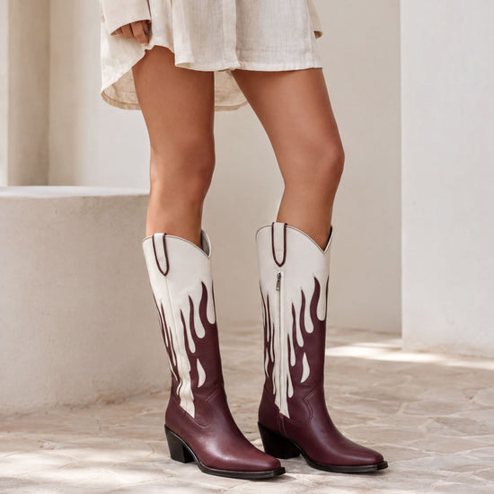 A person wearing a white dress and burgundy cowboy boots with flame designs stands in front of a white wall.
