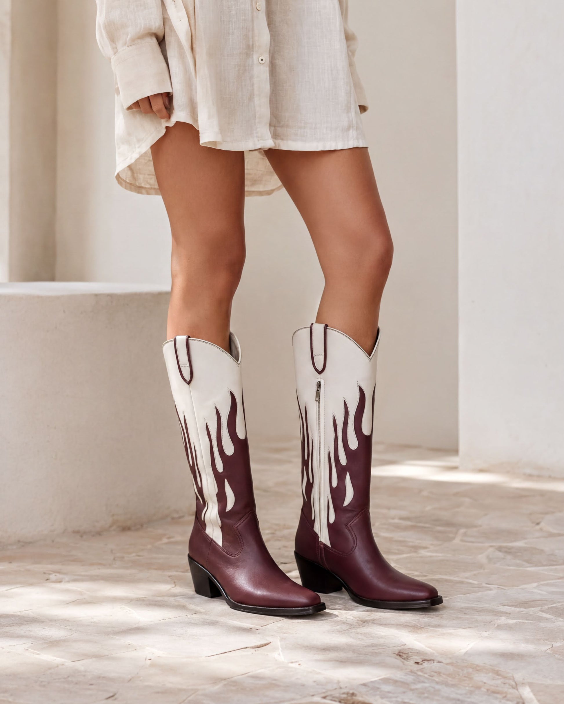 A person wearing a white dress and burgundy cowboy boots with flame designs stands in front of a white wall.
