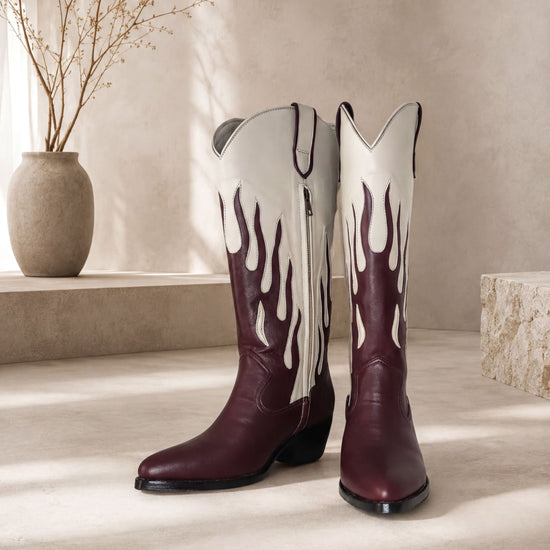A pair of burgundy cowboy boots with flame designs on the sides, placed on a light-colored surface against a beige wall.