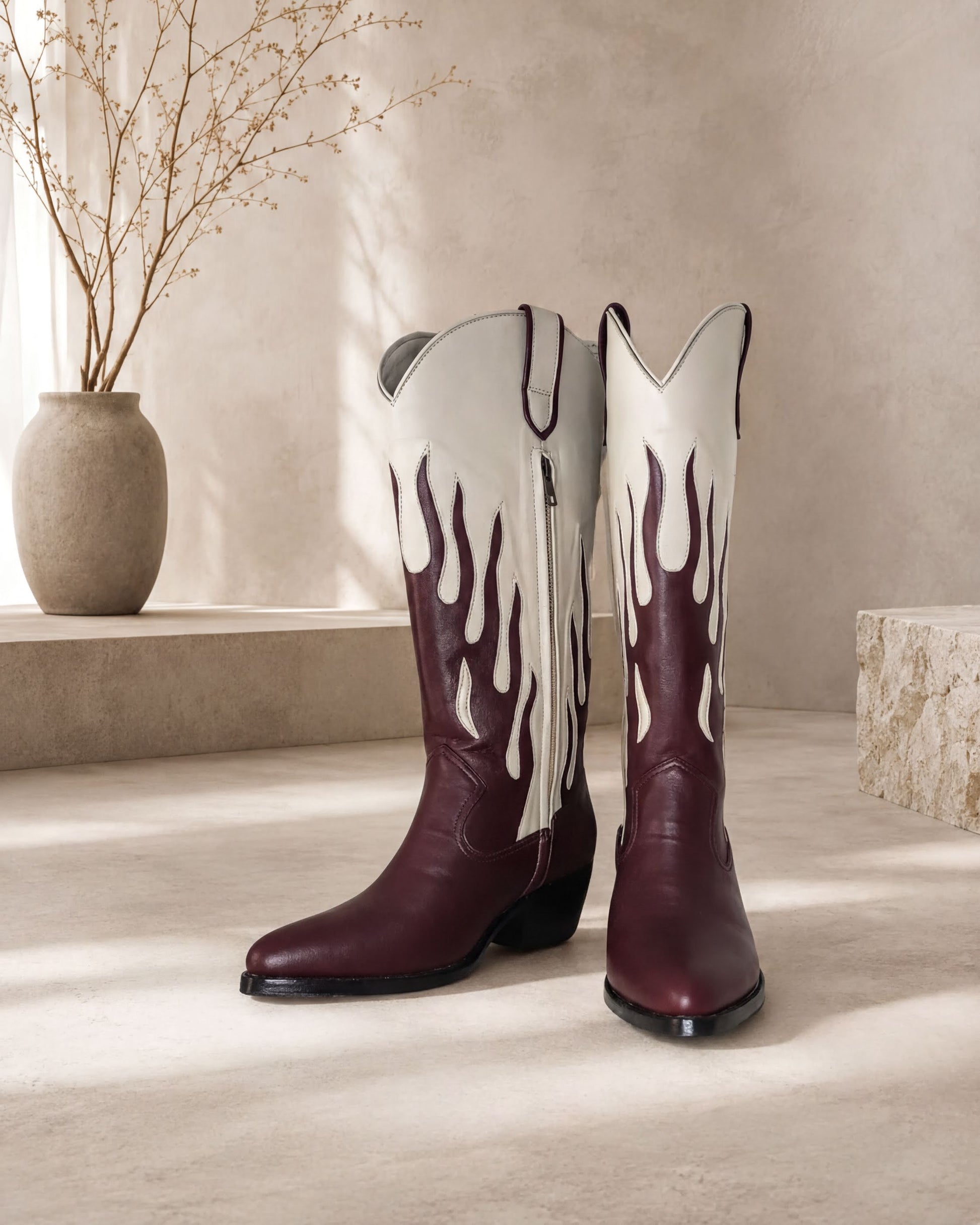 A pair of burgundy cowboy boots with flame designs on the sides, placed on a light-colored surface against a beige wall.