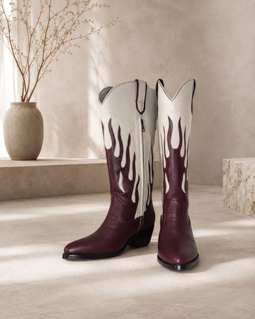 A pair of burgundy cowboy boots with flame designs on the sides, placed on a light-colored surface against a beige wall.