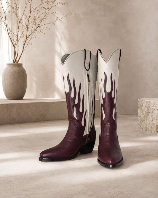 A pair of burgundy cowboy boots with flame designs on the sides, placed on a light-colored surface against a beige wall.