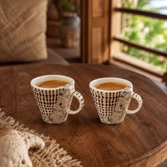 Two white coffee cups with a patterned design sit on a wooden table, with a beige pillow and a woven mat in the background.