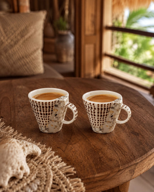 Two white coffee cups with a patterned design sit on a wooden table, with a beige pillow and a woven mat in the background.