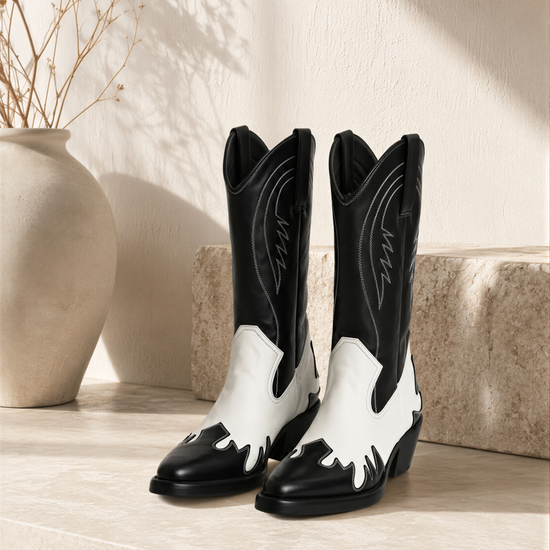 A pair of black and white cowboy boots is displayed on a white surface, with a vase of dried flowers and a stone wall in the background.