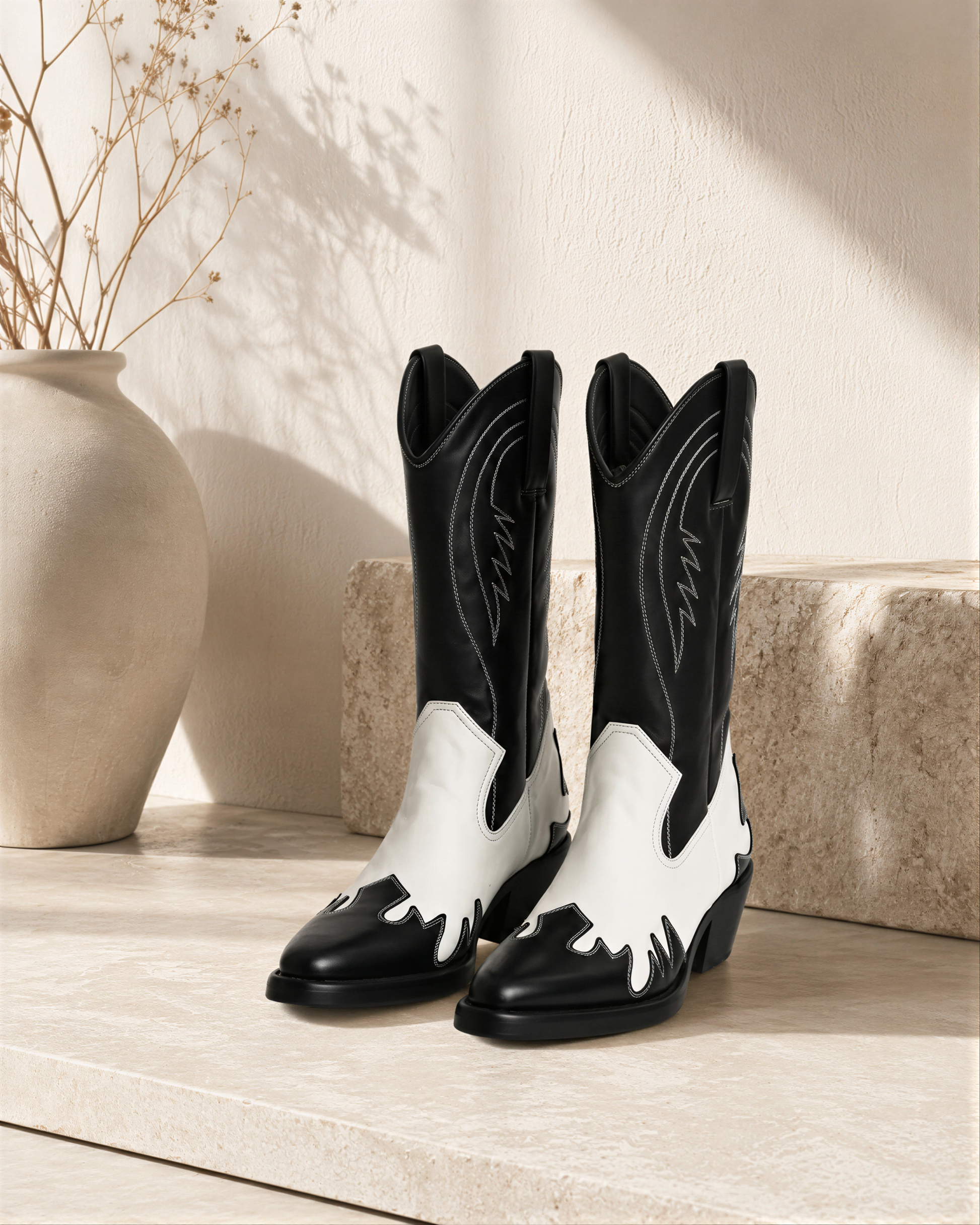 A pair of black and white cowboy boots is displayed on a white surface, with a vase of dried flowers and a stone wall in the background.