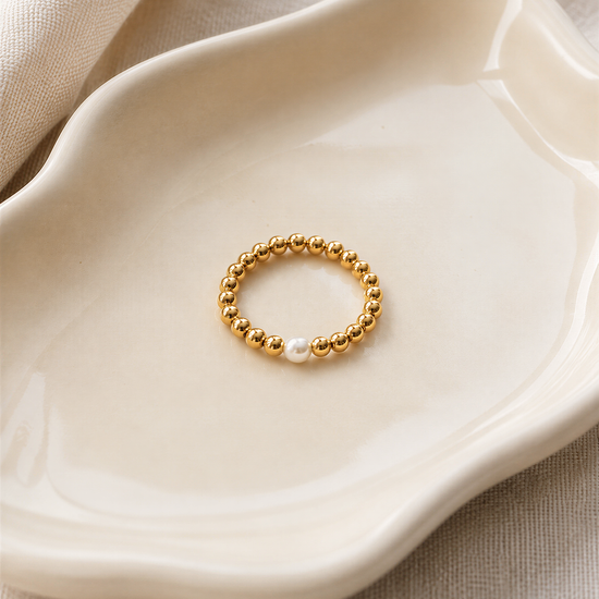 A gold-colored ring with a single pearl is displayed on a white ceramic plate.