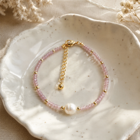 A delicate bracelet with pink beads and a pearl accent is displayed on a white plate, surrounded by a floral arrangement.