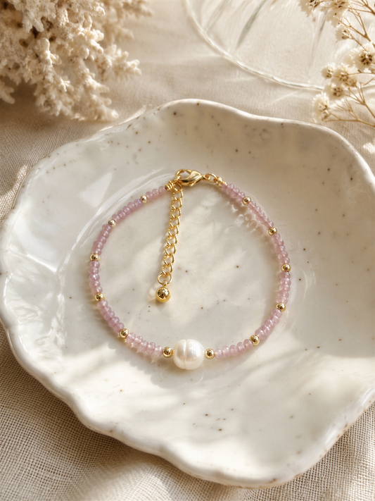 A delicate bracelet with pink beads and a pearl accent is displayed on a white plate, surrounded by a floral arrangement.