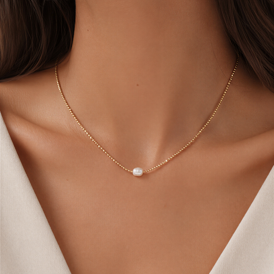 A close-up view of a woman's neck and chest area wearing a delicate gold necklace with a small white pearl pendant.