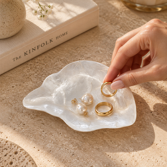 A hand is holding a pair of gold rings next to a white shell containing pearls and earrings, with a book titled "The Kinfolk Home" in the background.