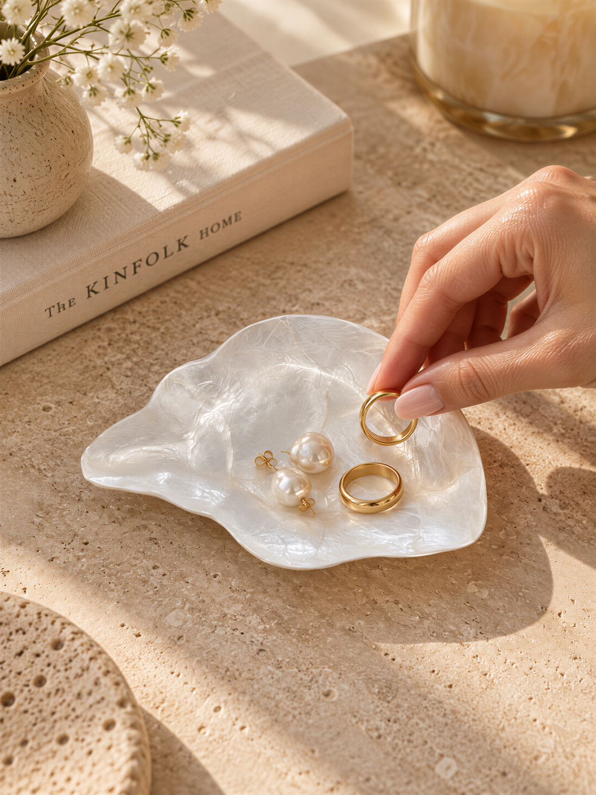 A hand is holding a pair of gold rings next to a white shell containing pearls and earrings, with a book titled "The Kinfolk Home" in the background.