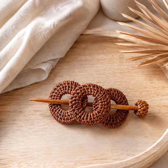 A woven hair accessory made of brown wicker circles rests on a wooden surface, accompanied by a white vase and a plant leaf.