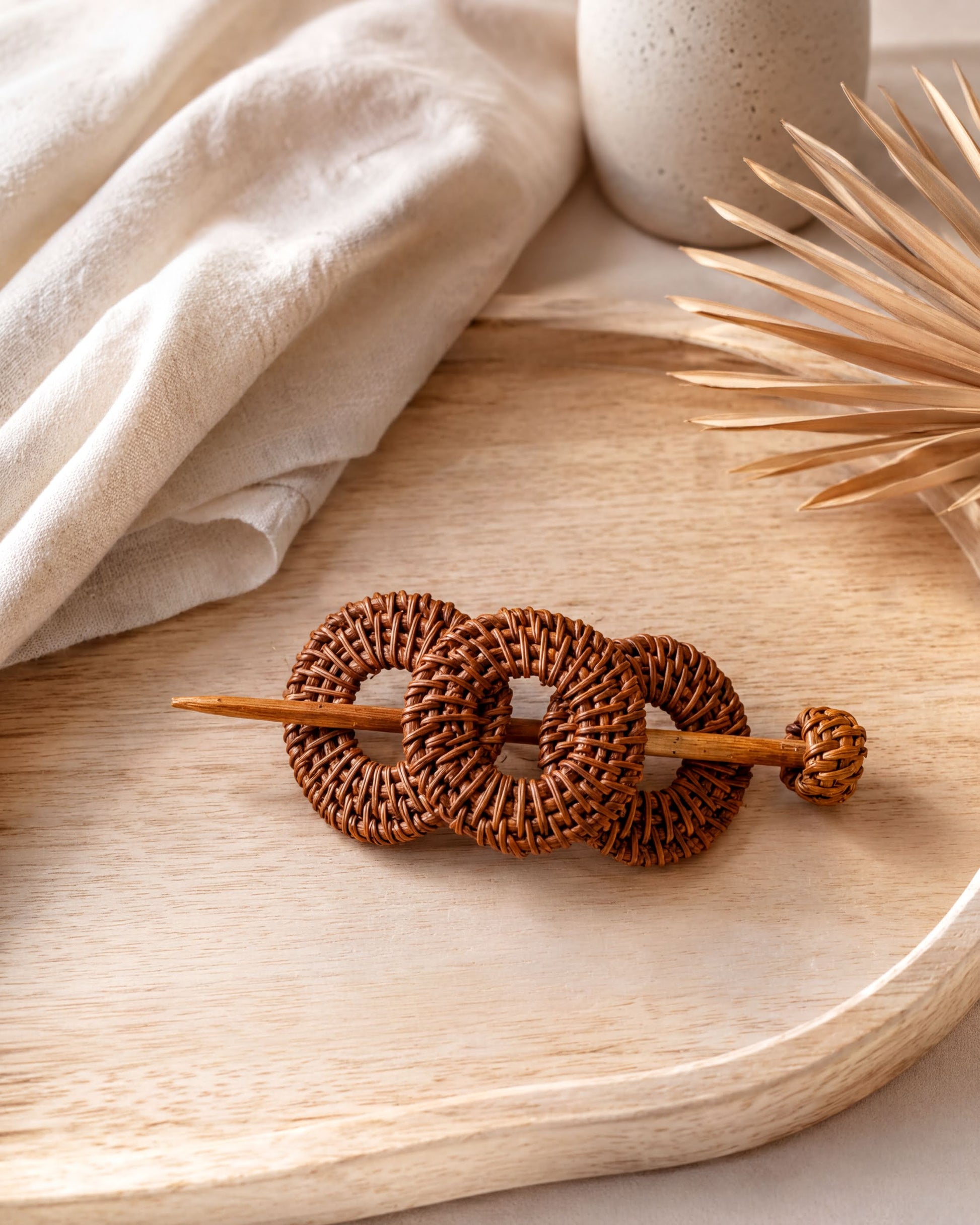 A woven hair accessory made of brown wicker circles rests on a wooden surface, accompanied by a white vase and a plant leaf.