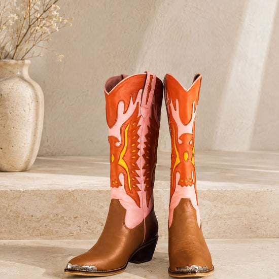 A pair of brown cowboy boots with vibrant orange and pink designs, placed on a white step against a plain background.