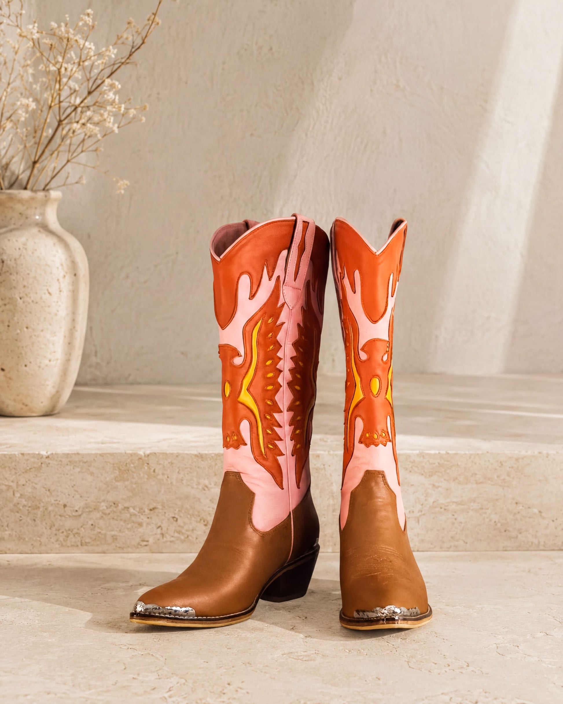 A pair of brown cowboy boots with vibrant orange and pink designs, placed on a white step against a plain background.