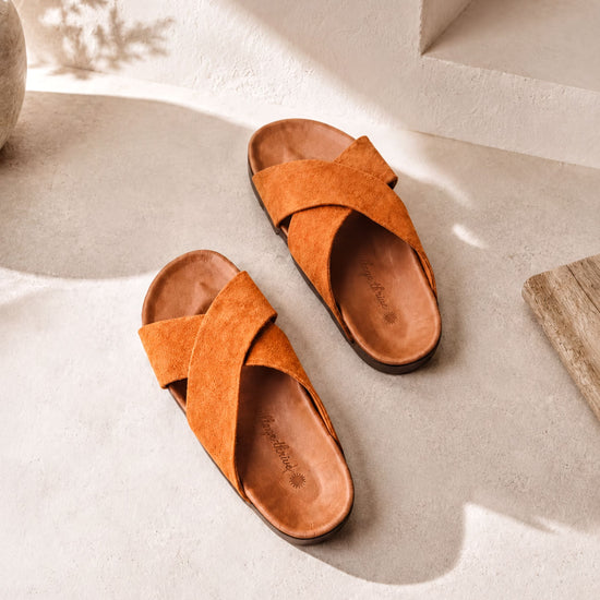 A pair of orange cross-strapped sandals is placed on a white surface, casting a shadow on the ground.