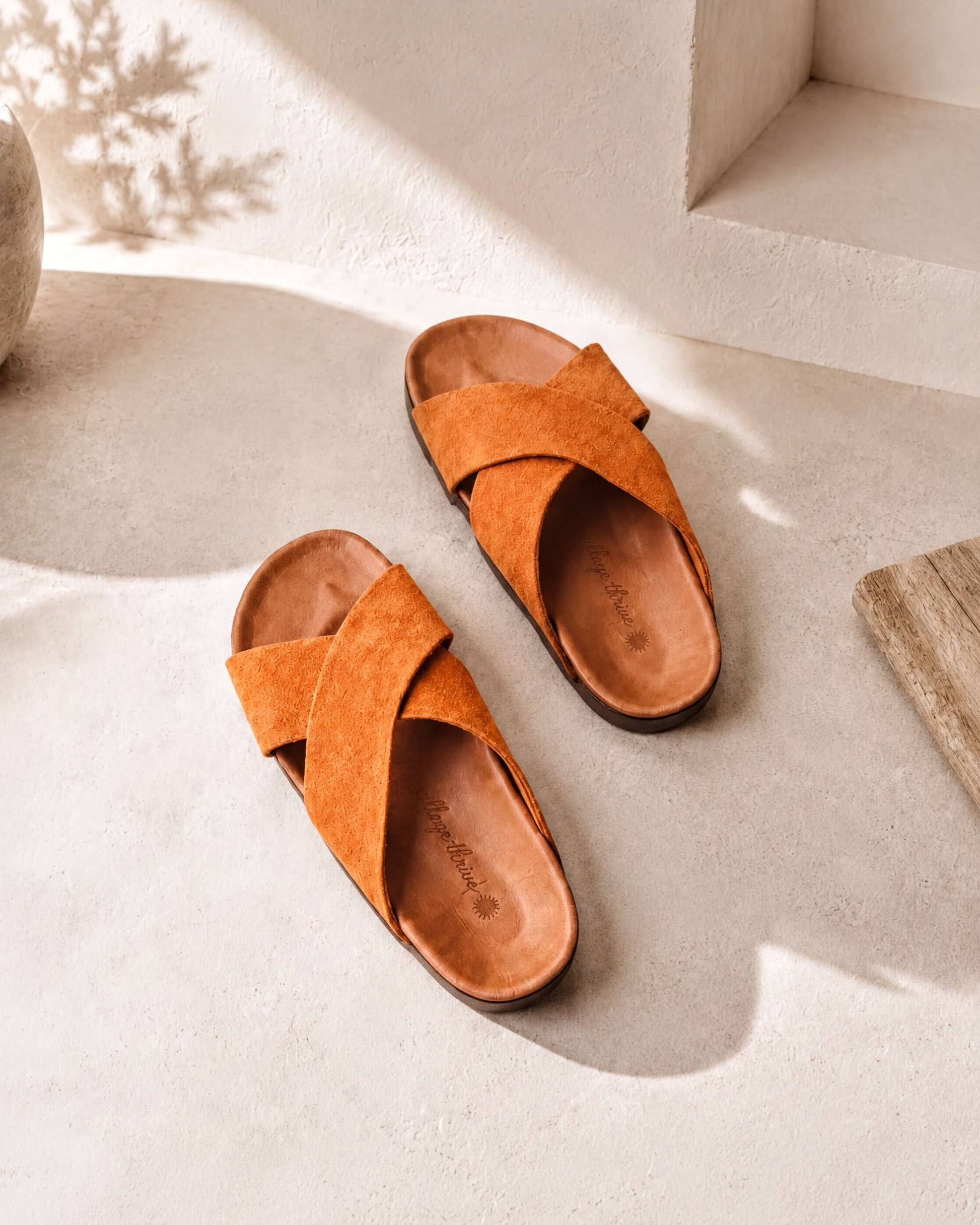 A pair of orange cross-strapped sandals is placed on a white surface, casting a shadow on the ground.