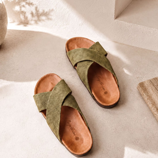 A pair of olive green cross-strapped sandals with a brown leather sole, placed on a white surface against a white wall.