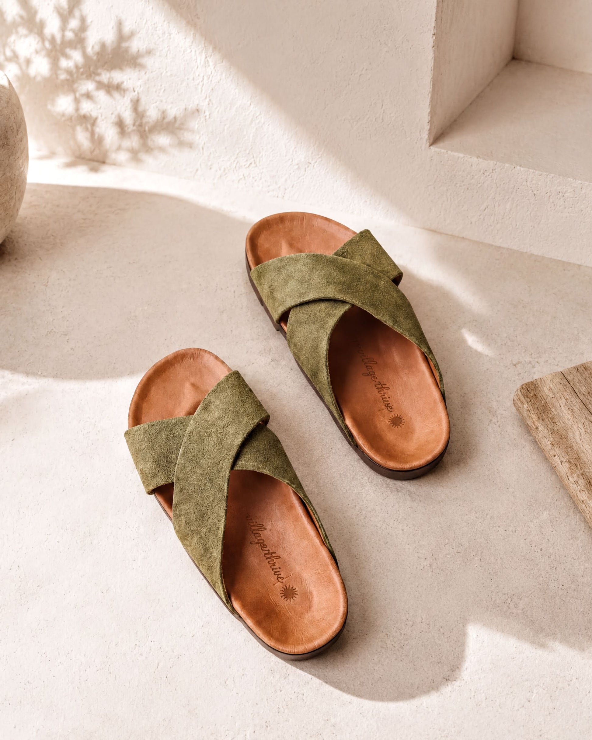 A pair of olive green cross-strapped sandals with a brown leather sole, placed on a white surface against a white wall.