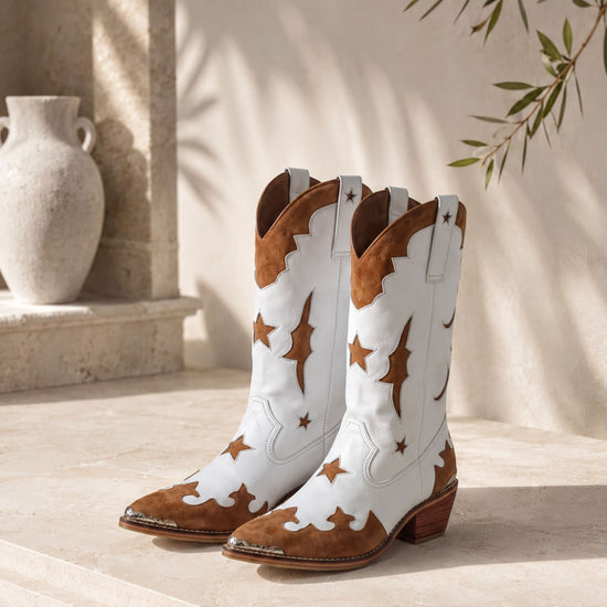 A pair of white cowboy boots with brown star and moon designs, placed on a stone floor against a beige wall with a plant.