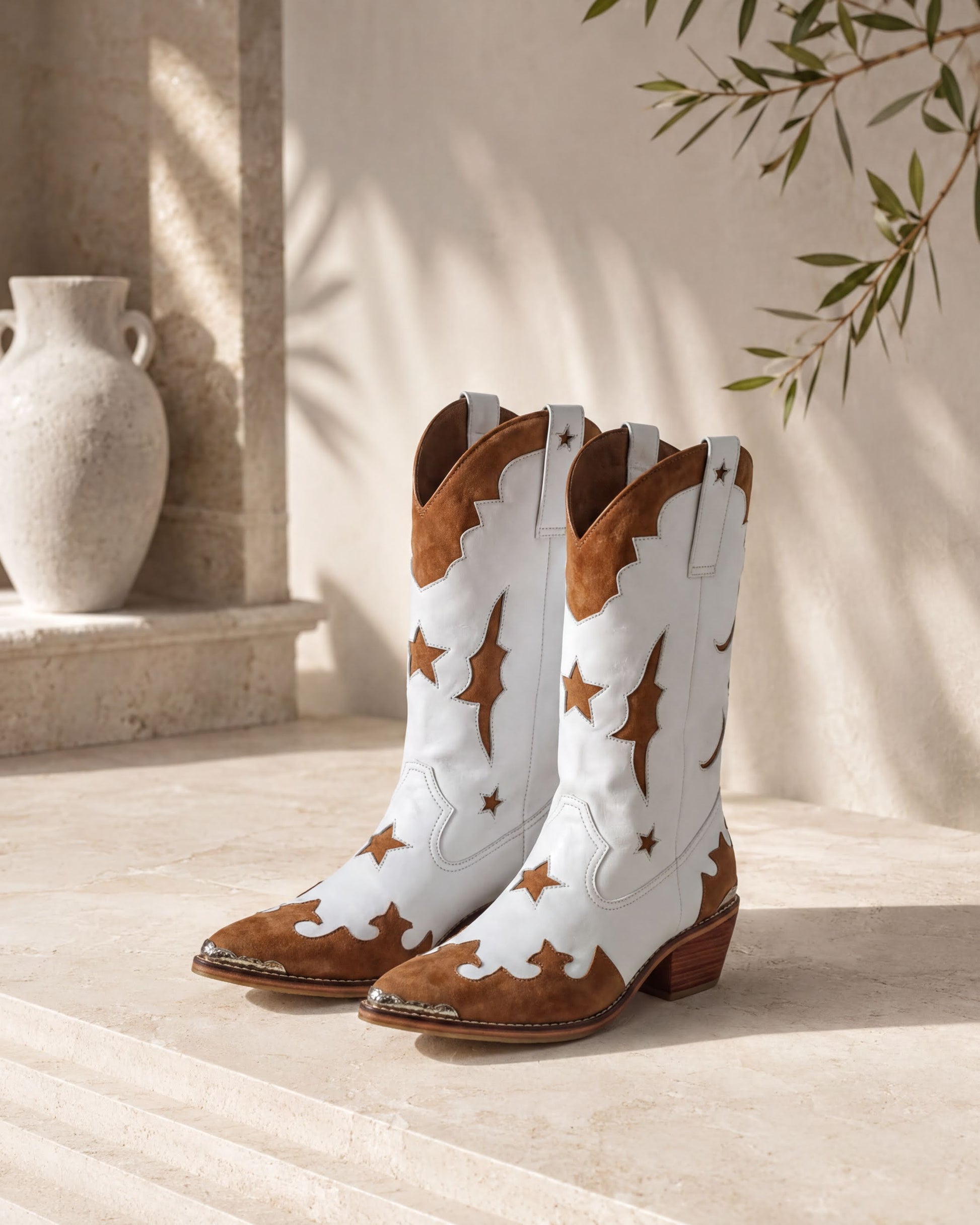 A pair of white cowboy boots with brown star and moon designs, placed on a stone floor against a beige wall with a plant.
