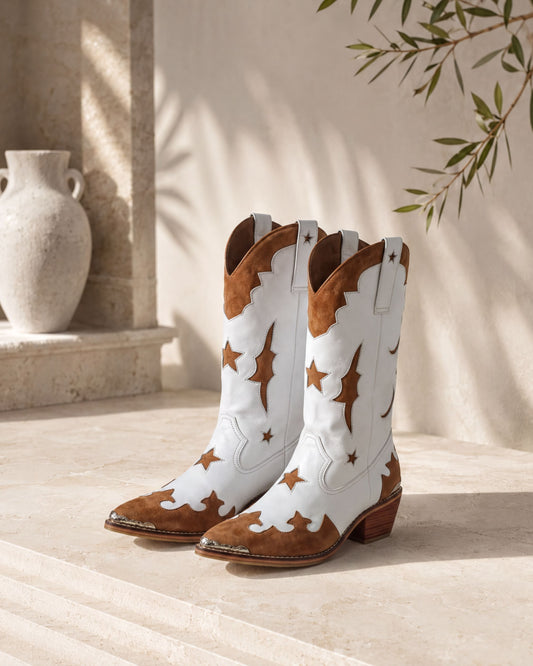 A pair of white cowboy boots with brown star and moon designs, placed on a stone floor against a beige wall with a plant.