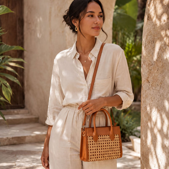 A woman in a white outfit stands in front of a stone archway, holding a woven purse.