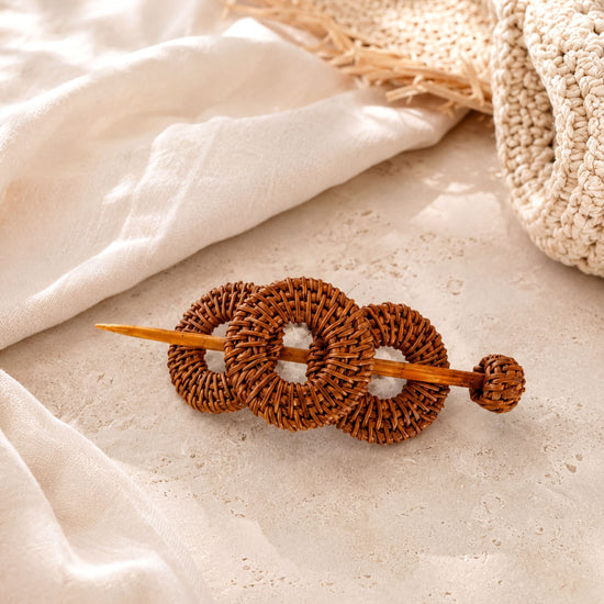 A woven hair accessory made of three circular wooden rings is placed on a white surface, with a woven straw hat visible in the background.