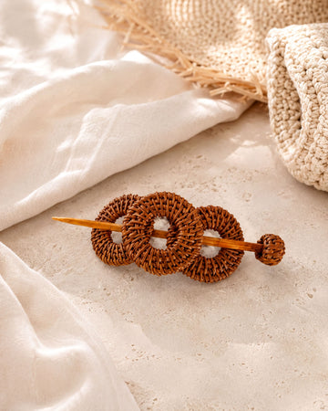A woven hair accessory made of three circular wooden rings is placed on a white surface, with a woven straw hat visible in the background.