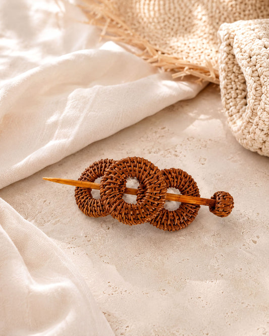 A woven hair accessory made of three circular wooden rings is placed on a white surface, with a woven straw hat visible in the background.